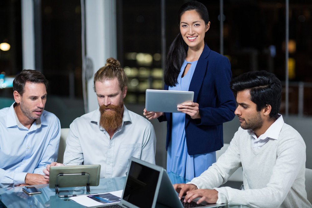 Businesswoman smiling at camera while colleagues discussing over digital tablet and laptop in the office