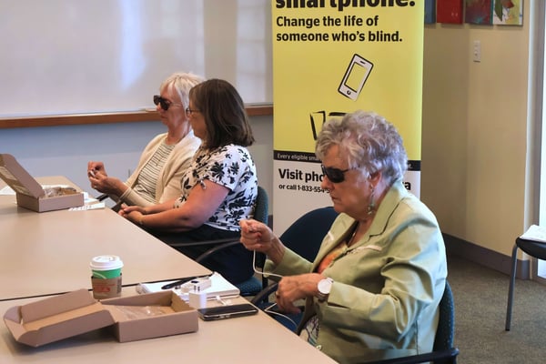 Two participants open smartphone boxes, with a Phone It Forward banner in the background.\